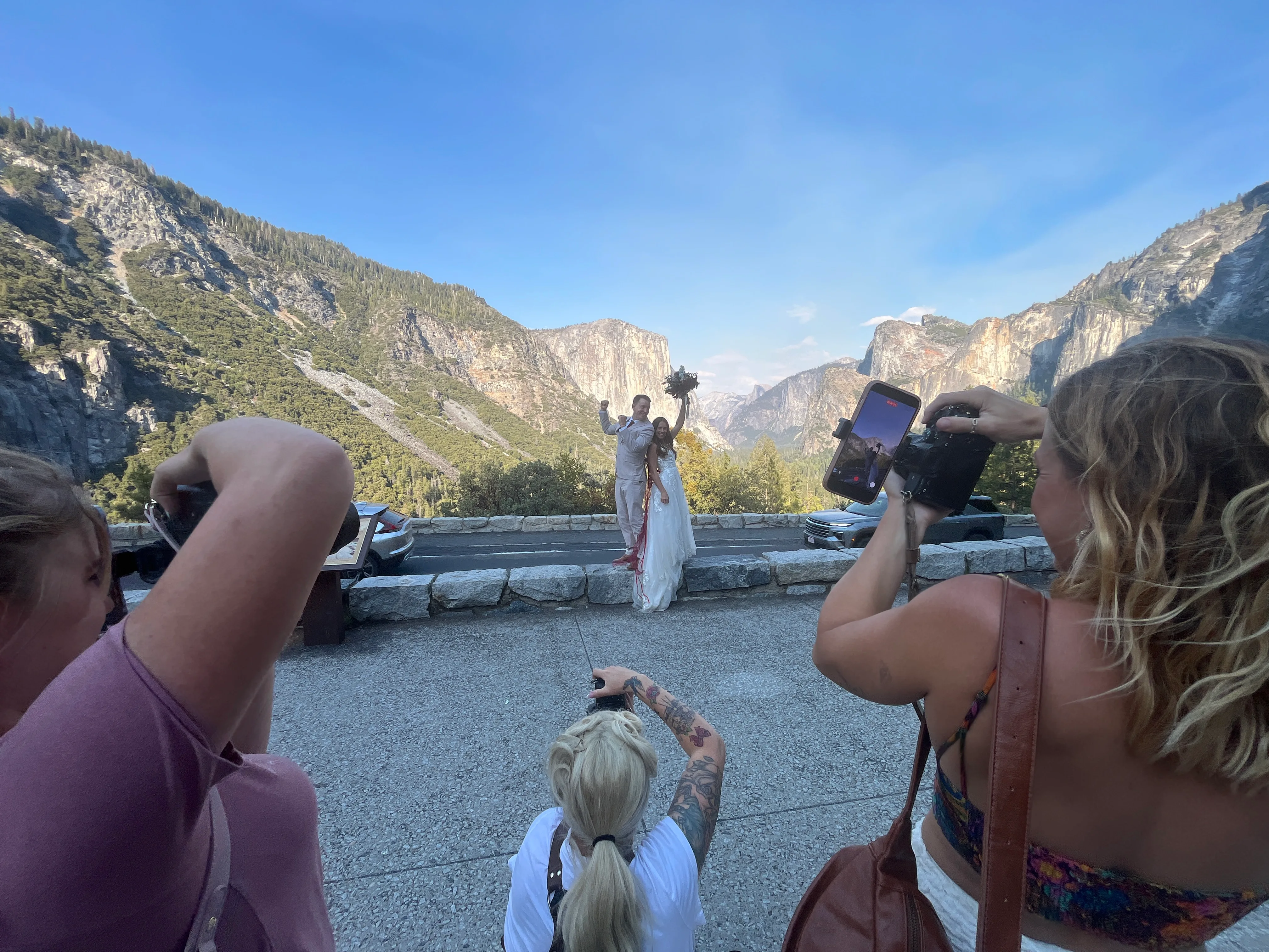 Photographers at the Yosemite tunnel parking lot shooting a bride and groom on a stone wall, the valley behind them.
