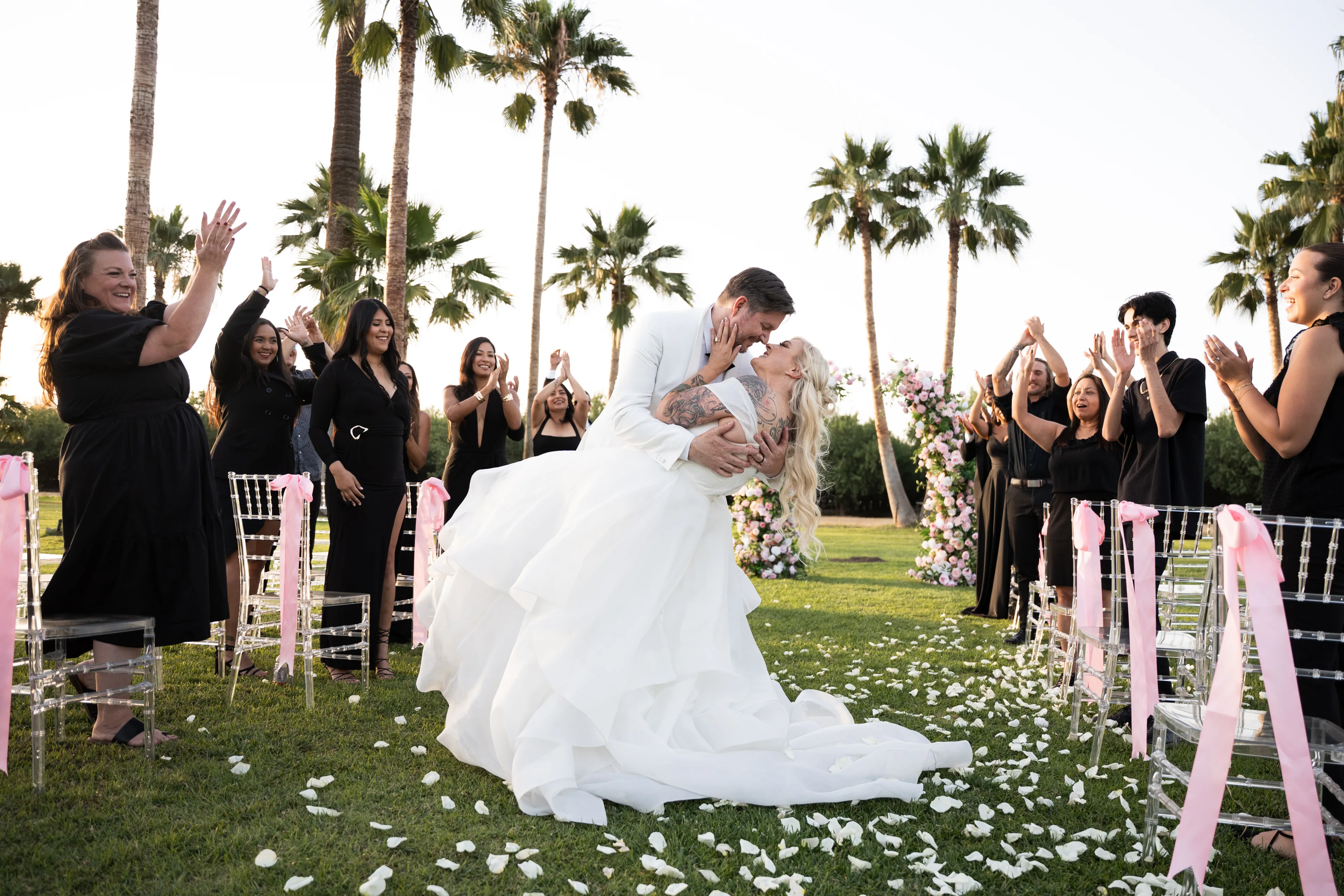Groom in white tuxedo dips a tattooed blonde bride in a layered gown as black-clad guests clap along a petal aisle.