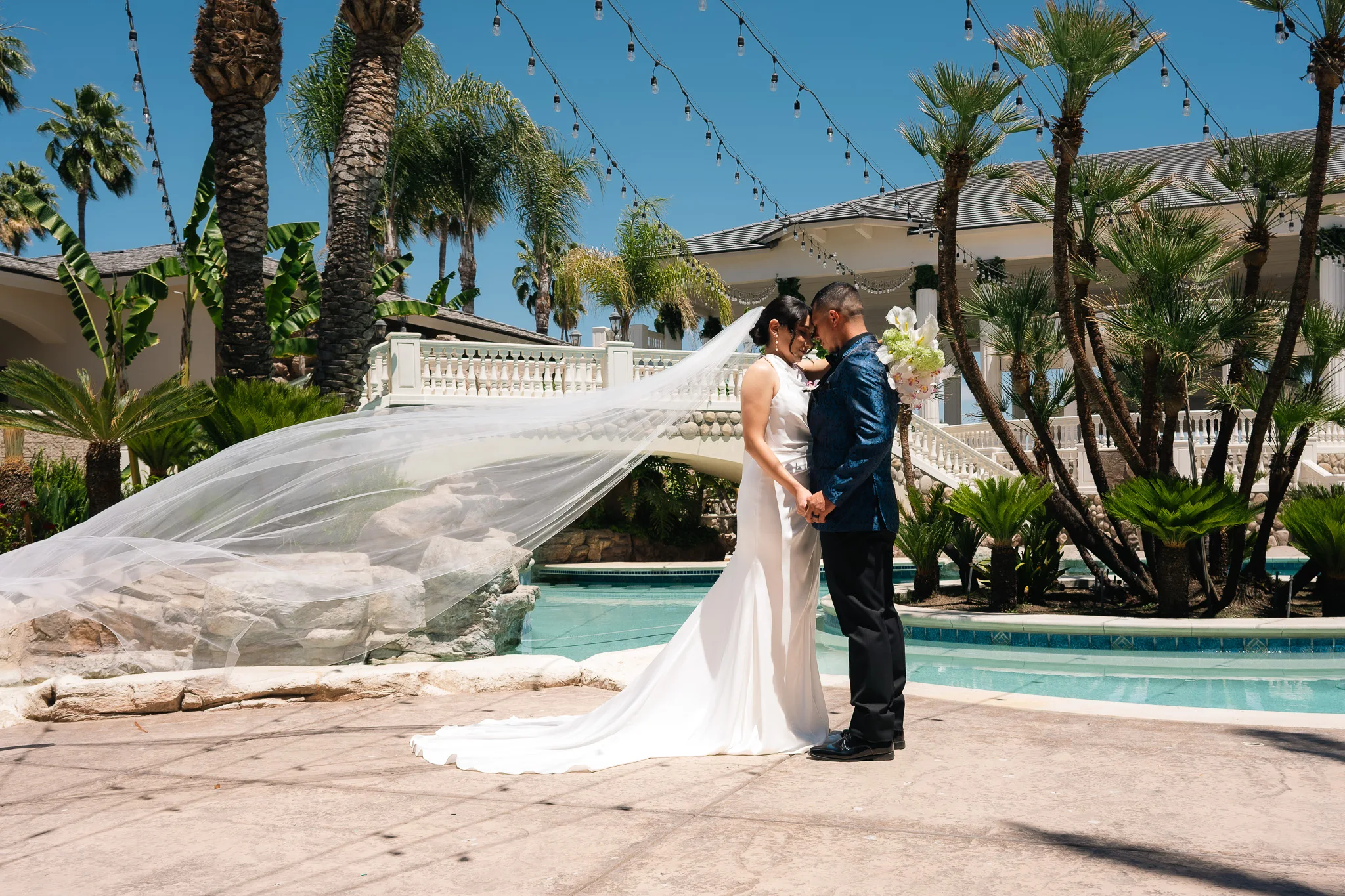 Bride in a white gown with a windblown veil stands forehead to forehead with a groom in a blue jacket by a pool.