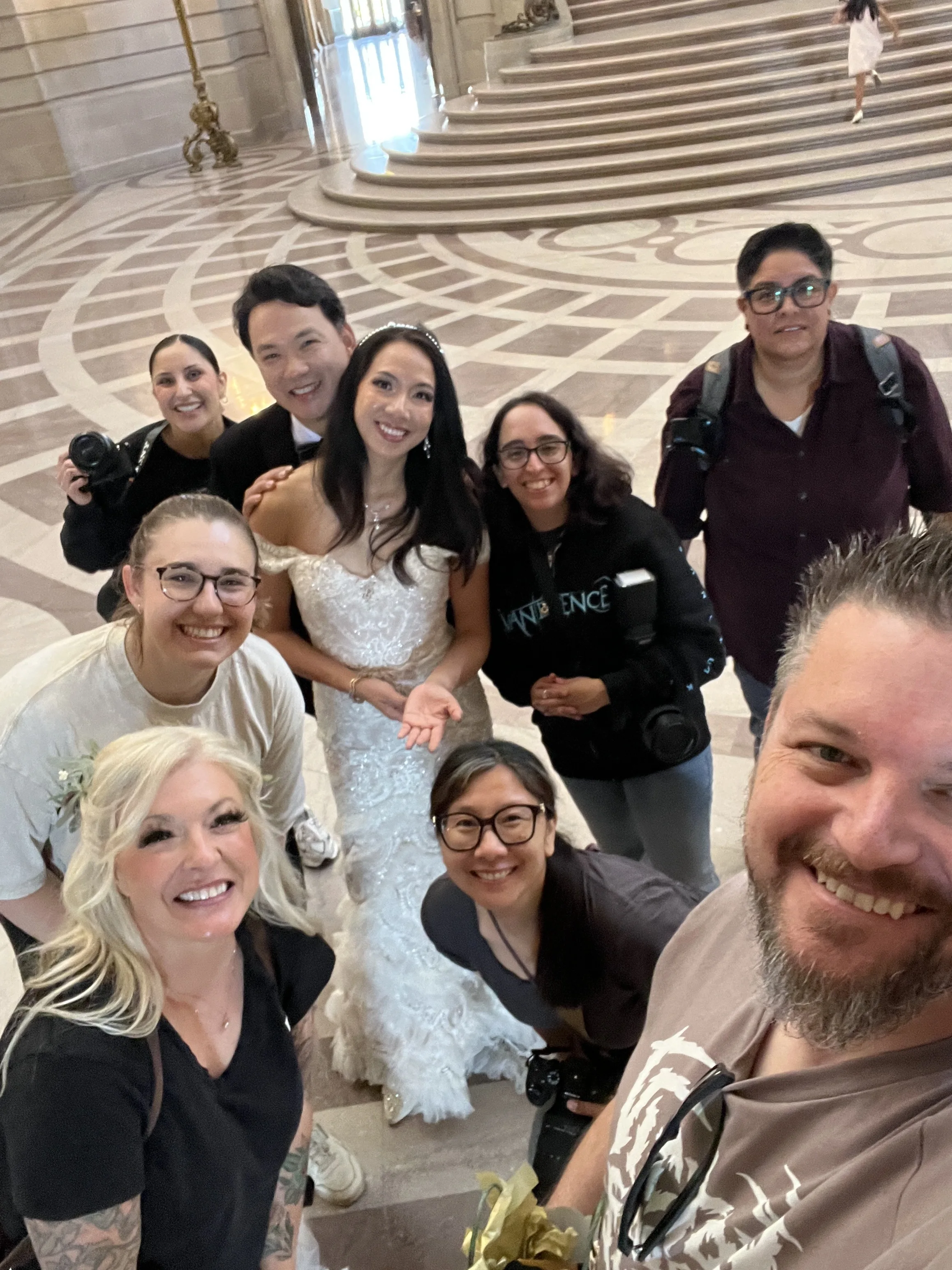Tilted group selfie of a bride in a white gown and a photo crew with cameras on a marble staircase.